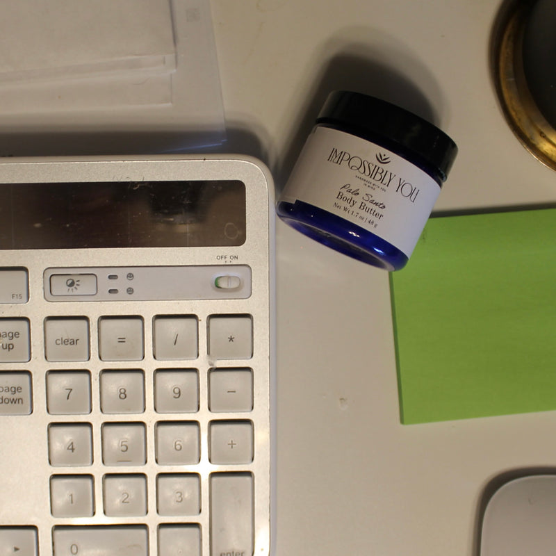 Travel size palo santo scented Impossibly You body butter next to a keyboard and post it notes on a desk.