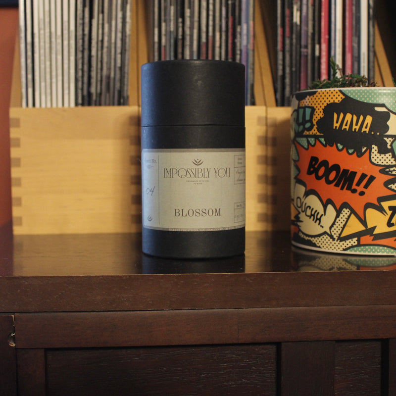 Blossom scented Impossibly You wax melts on a  shelf next a planter and magazines.